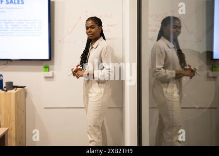 A young woman in white attire stands confidently presenting a sales strategy on a whiteboard in a modern coworking space, engaging her team members Stock Photo
