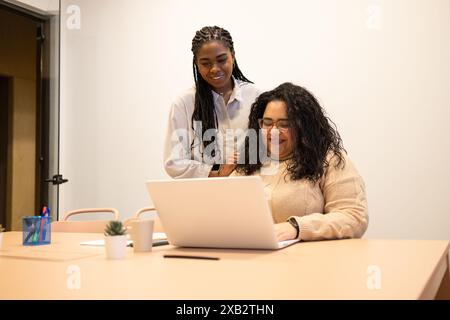 Two friends are engaged in a collaborative task at a coworking space, one standing and the other seated, both looking at a laptop screen with smiles, Stock Photo