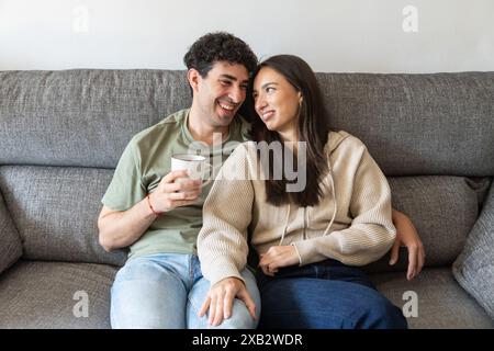 A happy young couple sitting comfortably on a gray sofa, enjoying time together with a cup of coffee Their smiles and interaction convey a warm, casua Stock Photo