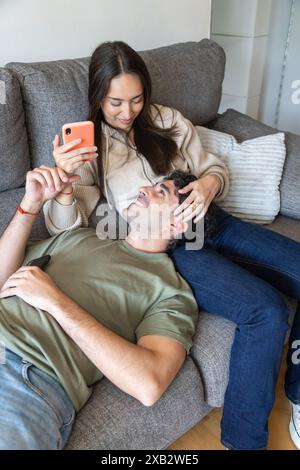 A young couple lounges comfortably on a gray sofa, sharing smiles while looking at a smartphone, depicting a cozy, casual moment at home Stock Photo