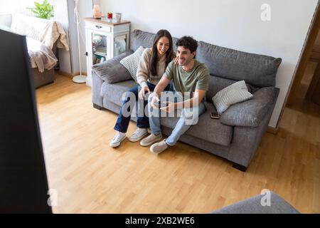 A young, cheerful couple enjoys a playful moment together while gaming on a console in their well-furnished, cozy living room, reflecting a relaxed an Stock Photo
