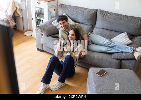 A young couple lounges comfortably in their living room, smiling and engaged in playing video games together, showcasing a moment of casual, joyful to Stock Photo