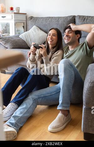 A joyful young couple sits on the floor, indulging in a lively video game Both are laughing and seem thoroughly engaged in their game, illustrating a Stock Photo