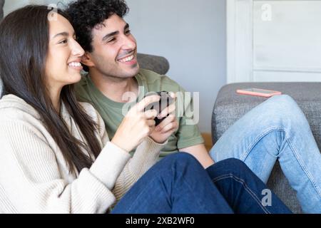 A cheerful couple sits on a couch playing video games, sharing joyful moments in a cozy living room setting, exemplifying content and relaxed leisure Stock Photo