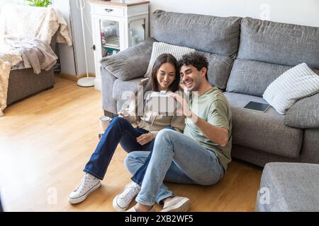 A young, cheerful couple sits on the floor at home, engaged in using a virtual reality headset, smiling and exploring digital worlds together, with a Stock Photo