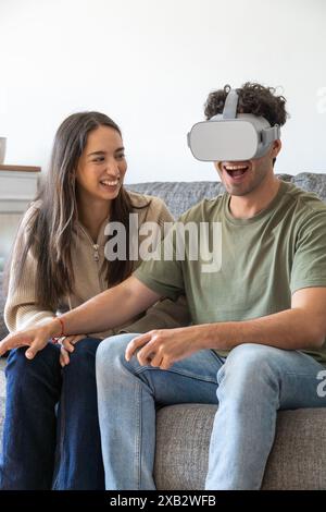 A jovial couple sits on a sofa while the man, wearing a virtual reality headset, reacts animatedly to his experience The woman, amused, looks on with Stock Photo