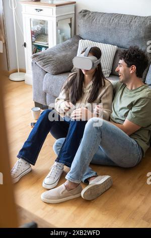 A young couple sits on the living room floor, with the woman wearing a virtual reality headset and the man smiling playfully beside her, suggesting a Stock Photo