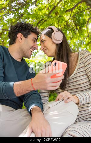 A young couple enjoys a sunny day outdoors, sharing smiles and looking at a smartphone together, conveying a sense of happiness and connectivity with Stock Photo