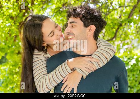 An affectionate couple shares a tender moment, with the woman lovingly embracing the man from behind, both smiling joyfully in a lush green park Stock Photo