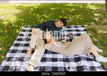 A young man and woman enjoy a peaceful moment on a black and white checkered blanket in a sunny park, browsing a smartphone together Stock Photo