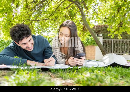 A joyful young couple enjoys a relaxing day in the park, studying and using a smartphone under the shade of green trees Their casual clothes and seren Stock Photo