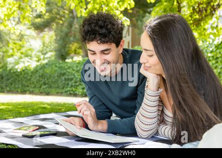 A young couple shares a joyous moment reading together on a picnic blanket under the shade of lush green trees in a serene park Stock Photo