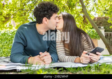 A young couple enjoys a romantic moment, sharing a kiss while lounging on a blanket in a lush garden The man kisses his partner affectionately as she Stock Photo