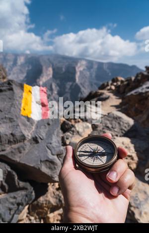 Anonymous person holds a compass while exploring rocky terrain in Oman, with a trail marker visible and mountains in the background Stock Photo