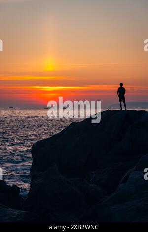 Silhouette of an unrecognizable individual stands atop a rocky outcrop against the vibrant backdrop of a sunset over the ocean near the Atlanterra Bea Stock Photo