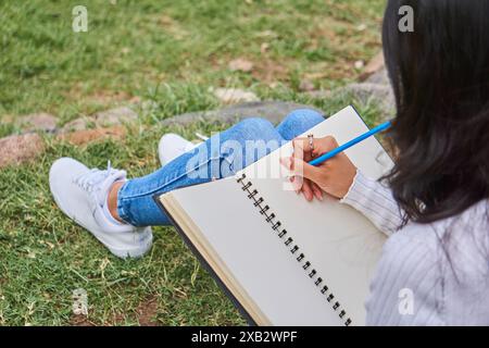 A woman enjoys a peaceful moment in the park, jotting down thoughts in a spiral notebook while reclining on the grass Stock Photo