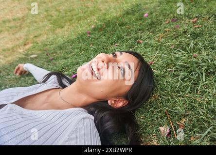 A cheerful young woman lies on the grass, smiling brightly as she relaxes in a serene park setting Stock Photo