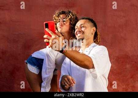 A joyful curly-haired woman and a smiling man with dreadlocks are capturing a selfie moment together using a red smartphone Both are dressed in casual Stock Photo
