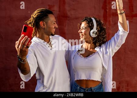 A joyful young couple standing against a red wall, the man taking a selfie with a red smartphone and the woman wearing headphones and dancing Stock Photo