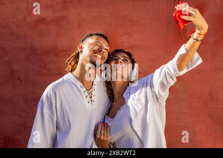 A young, joyful couple, both dressed in white shirts, laugh and take a selfie together against a vibrant red wall The woman is using headphones and a Stock Photo