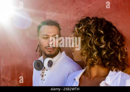 A young man with dreadlocks and headphones hanging from his neck exchanges an affectionate gaze with a young woman with curly hair Both are dressed in Stock Photo