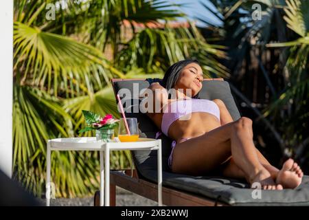 A Latin woman enjoys sunshine while reclining on a sunbed, eyes closed, in tropical ambiance. Stock Photo