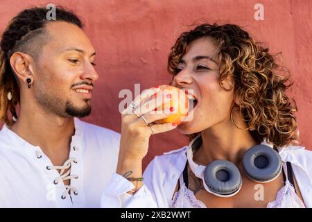 A young woman with curly hair bites into a red apple, sharing a joyful moment with a smiling young man beside her against a soft pink background Stock Photo