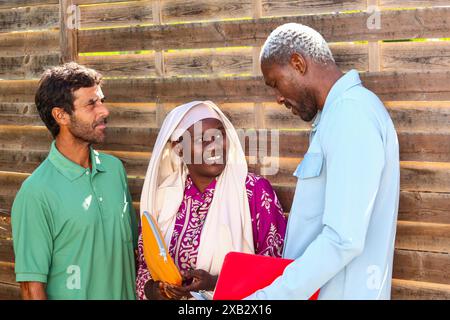 Three people having a casual outdoor meeting, smiling and sharing documents, with wooden fencing and greenery in the background, symbolizing a summer Stock Photo