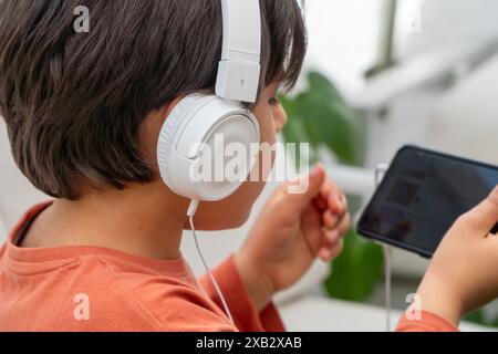A child in a casual orange shirt listens attentively to music through white over-ear headphones, holding a black tablet in a bright, indoor setting Stock Photo