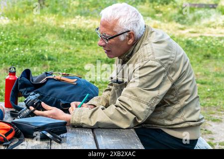 Senior man with white hair sits outdoors at a wooden table, examining a camera and taking notes, with personal items and a lush green background Stock Photo