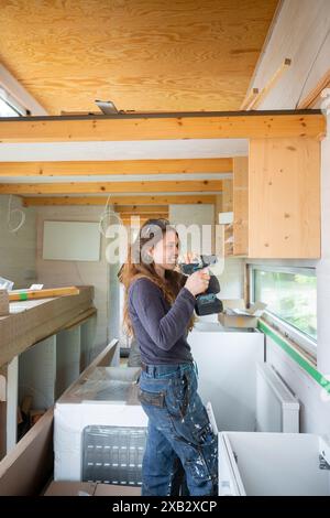 A young woman, casually dressed in work clothes, is smiling while using a cordless drill in a sunlit wooden workshop, working on her DIY project. Stock Photo