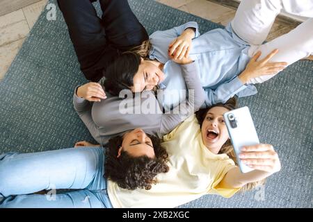 Top view of three friends lying on the floor at home, joyfully taking a group selfie, capturing a casual and cheerful moment. Stock Photo