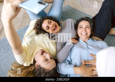 From above three female friends lying on the floor, sharing a joyful moment as they take a selfie together, exuding happiness and camaraderie. Stock Photo