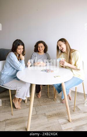 Three women share laughter and joy while sitting at a table playing cards, creating a warm, friendly atmosphere in a cozy home setting. Stock Photo