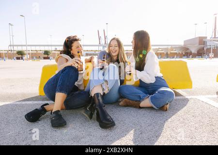 Three young women sit by the dockside, sharing laughter and drinks under the bright sun. Stock Photo