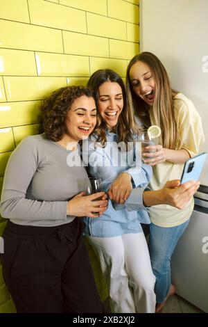 Three female friends look at a smartphone and laugh together, sharing a moment of joy with drinks in hand against a yellow-tiled background. Stock Photo