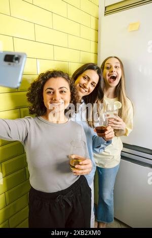 Three happy female friends take a selfie together at home, casually dressed, drinking and enjoying each other's company. Stock Photo
