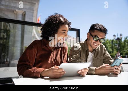 A Chinese man and a Hispanic woman enjoy a sunny day in Madrid, Spain, sitting at an outdoor table using their tablet and smartphone while smiling at Stock Photo