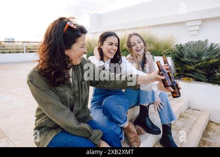 Three women share a cheerful moment while drinking beer together in a port area, chatting and laughing. Stock Photo