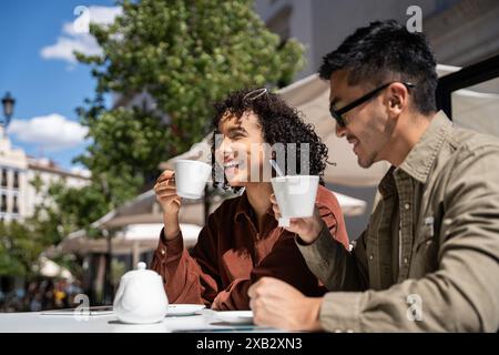 A multiethnic couple, a Chinese man, and a Hispanic woman, smile and enjoy coffee at an outdoor café in Madrid, Spain, on a sunny day. The background Stock Photo