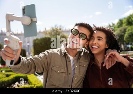 A multiethnic couple, a Chinese man, and a Hispanic woman, are taking a selfie with a smartphone on a gimbal while enjoying a sunny day in a beautiful Stock Photo