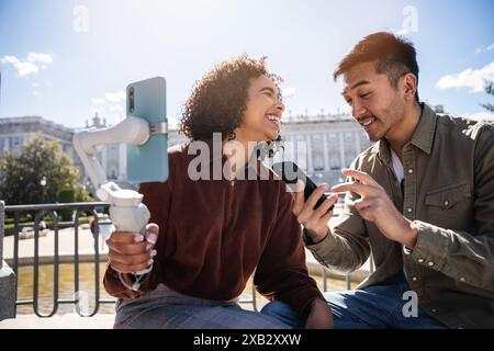 A cheerful Chinese man and Hispanic woman enjoy a sunny day in Madrid, Spain. They are smiling and using their smartphones to capture the moment in fr Stock Photo