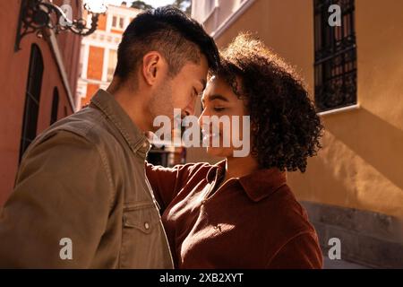 A Chinese man and a Hispanic woman share a tender moment on a picturesque street in Madrid, Spain, under the warm, golden sunlight. Their smiles and t Stock Photo