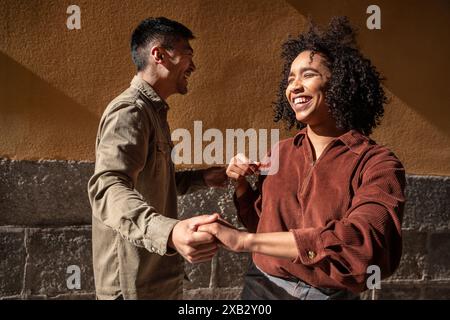 A Chinese man and a Hispanic woman share a laugh while holding hands in Madrid, Spain. The couple is enjoying a sunny day, reflecting cross-cultural c Stock Photo