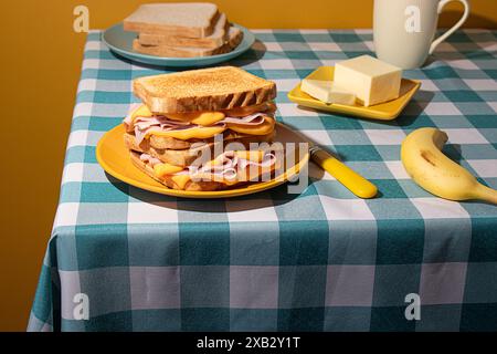 A homemade breakfast on a checkered blue tablecloth features a sandwich, banana, coffee, and butter against yellow background Stock Photo