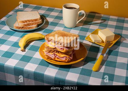 Sandwich on a yellow plate accompanies a cup of coffee, butter on a small plate, and a fresh banana, all arranged on a blue and white checkered blue t Stock Photo