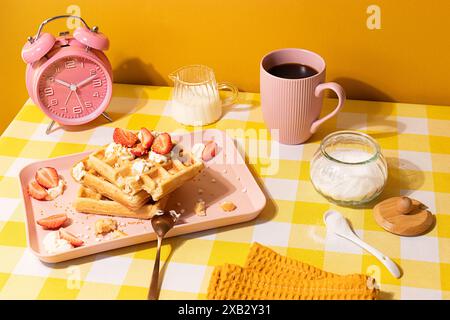 Colorful breakfast scene featuring waffles topped with strawberries and whipped cream, served on a pastel pink plate with coffee and milk on a yellow Stock Photo