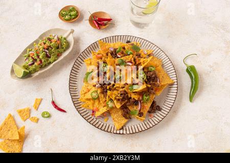 Top view of a platter of nachos generously topped with cheese, ground beef, sliced jalapenos, and fresh tomato, served alongside guacamole and a lime Stock Photo