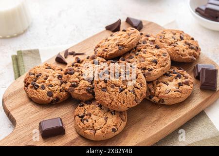 Freshly baked chocolate chip cookies arranged on a wooden cutting board, accompanied by milk and chocolate pieces Stock Photo