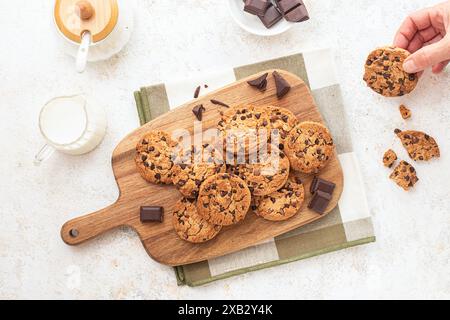 A delightful display of freshly baked chocolate chip cookies arranged on a wooden cutting board with dark chocolate pieces and a cup of milk on the si Stock Photo